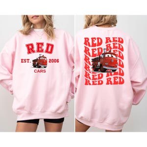 May include: Pink sweatshirt with a red graphic of a fire truck and the text "RED EST. 2006 CARS" on the front. The back of the sweatshirt has the word "RED" repeated in a diagonal pattern.