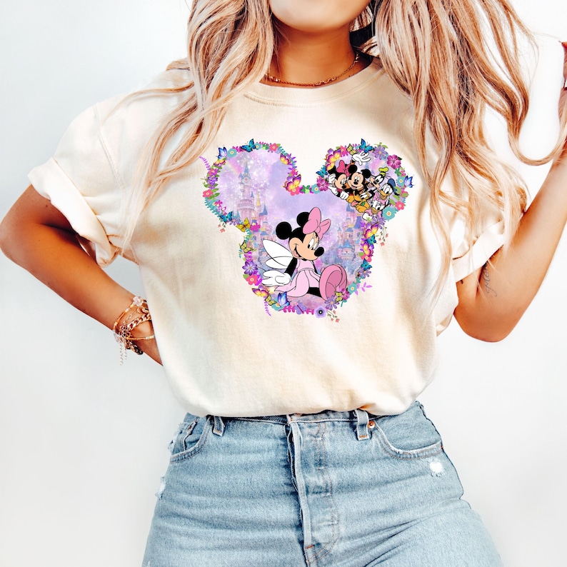 May include: A white t-shirt with a colorful graphic of Mickey Mouse's head. The graphic features Minnie Mouse in a pink dress and wings, surrounded by flowers and a purple castle.