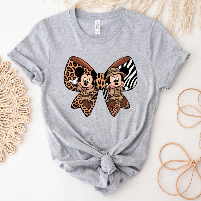 May include: A light grey t-shirt with a large bow design featuring Mickey and Minnie Mouse characters. The bow has a leopard print and zebra stripe pattern. Mickey and Minnie are in safari outfits.