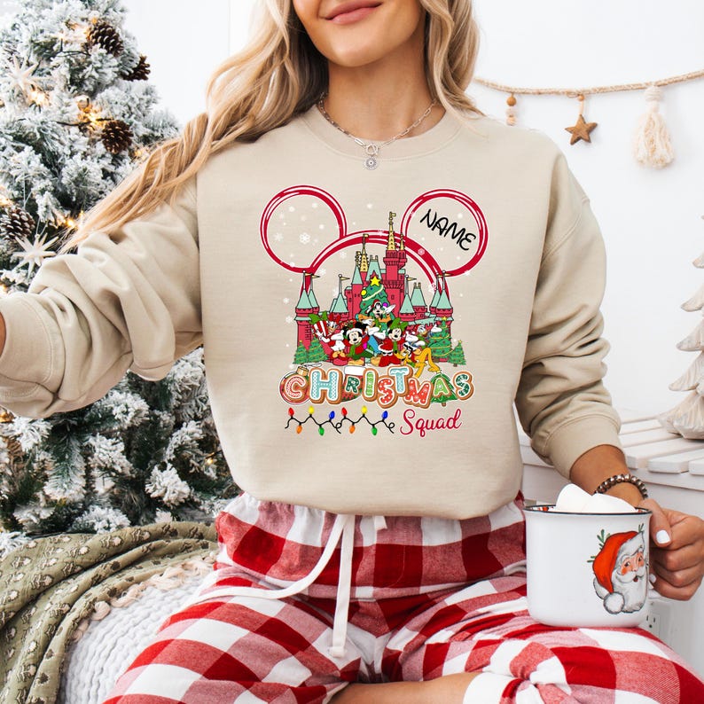 Puede incluir: Sudadera beige con un gr&aacute;fico colorido de un castillo, personajes de Disney y el texto "Christmas Squad". El dise&ntilde;o incluye orejas de Mickey Mouse y un espacio para un nombre. Una cadena de luces navide&ntilde;as est&aacute; en la parte inferior.