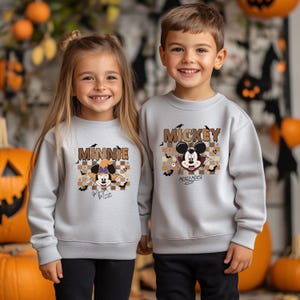 May include: Two children wearing matching gray sweatshirts with Halloween-themed Disney character designs. One sweatshirt says "Minnie" and features Minnie Mouse in a witch hat. The other says "Mickey" and features Mickey Mouse in a vampire costume.