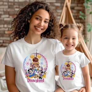 Op de afbeelding: Twee witte T-shirts met een kleurrijke afbeelding van Mickey Mouse en zijn vrienden die het 70-jarig jubileum van Disneyland vieren. De afbeelding bevat vuurwerk, een kasteel en de tekst "Disneyland Resort".