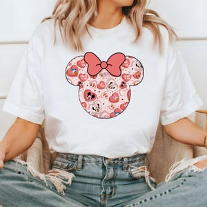 May include: White t-shirt with a pink and red Minnie Mouse head graphic. The graphic features a pink bow and a pattern of hearts and cartoon characters.