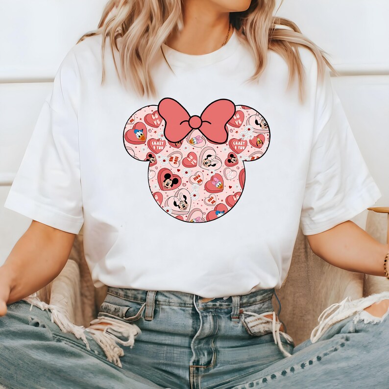 May include: White t-shirt with a pink and red Minnie Mouse head graphic. The graphic features a pink bow and a pattern of hearts and cartoon characters.