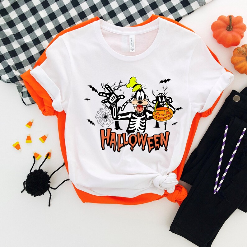 May include: White Halloween-themed t-shirt featuring a cartoon character in a skeleton costume. The design includes the word "Halloween" in orange, bats, and a spiderweb. The shirt has orange sleeves and is paired with black pants and Halloween decorations.