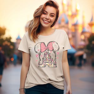 May include: A beige t-shirt with a pink bow and a cartoon drawing of a castle with fireworks. The castle is surrounded by hearts and balloons.