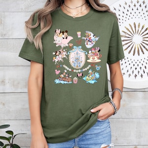 May include: Olive green t-shirt featuring a collage of cartoon characters, including Mickey and Minnie Mouse, with a castle and the text "Best Day Ever." The design includes various scenes and elements in a colorful, whimsical style.