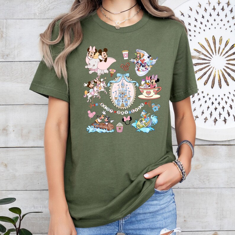 May include: Olive green t-shirt featuring a collage of cartoon characters, including Mickey and Minnie Mouse, with a castle and the text "Best Day Ever." The design includes various scenes and elements in a colorful, whimsical style.