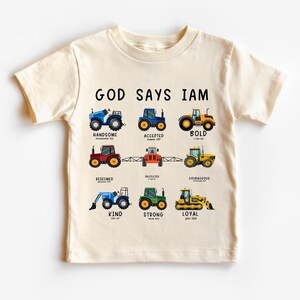 May include: A beige t-shirt with the text "GOD SAYS IAM" printed on the front. The text is surrounded by illustrations of tractors with different colors and designs. Each tractor is labeled with a positive word, such as "handsome", "accepted", "bold", "redeemed", "protected", "courageous", "kind", "strong", and "loyal".