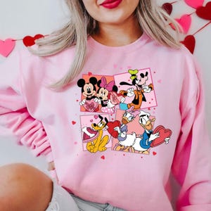 May include: Pink sweatshirt with a graphic print of Mickey Mouse, Minnie Mouse, Goofy, Pluto, and Donald Duck holding Valentine's Day hearts.