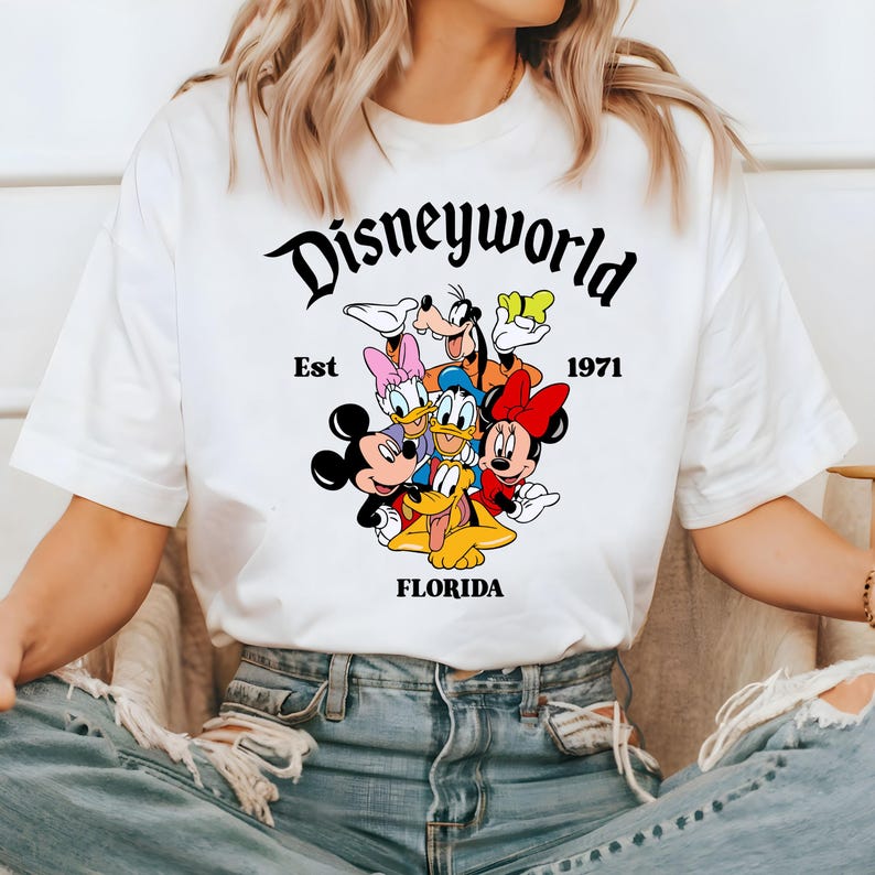 Pu&ograve; includere: Maglietta bianca con una grafica nera di Topolino, Minnie Mouse, Paperino, Pippo e Pluto. La grafica include il testo "Disneyworld Est 1971 Florida".