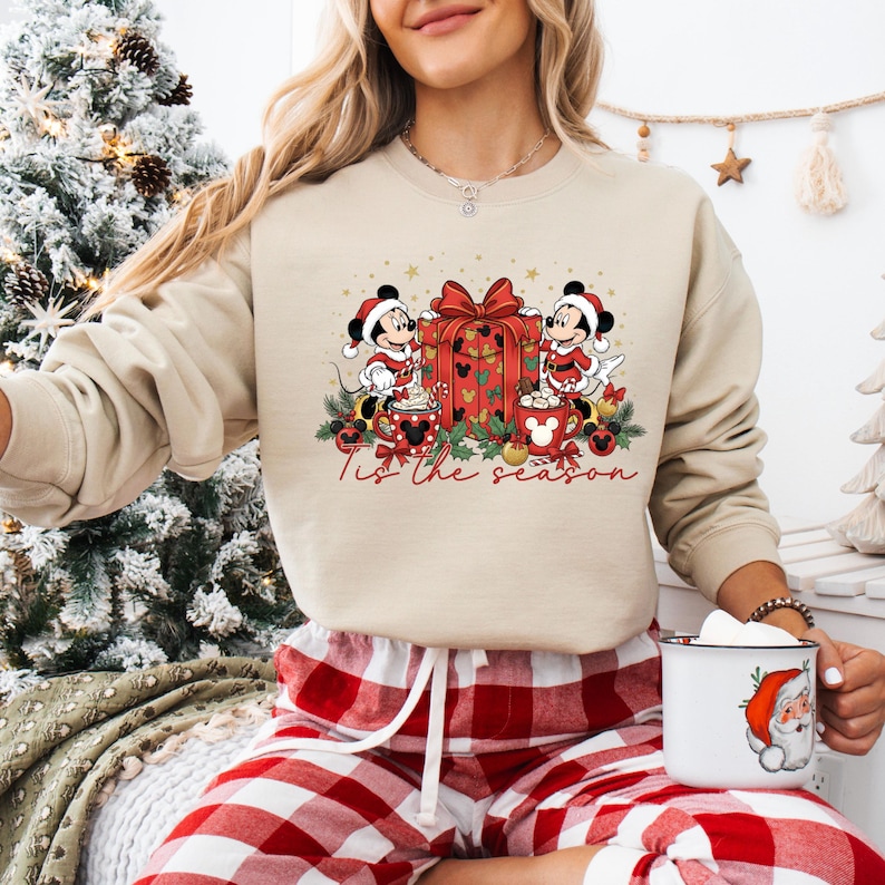 May include: Beige sweatshirt featuring a festive graphic with Mickey and Minnie Mouse in Santa hats, a wrapped gift, and mugs of hot chocolate. The text "Tis the season" is written in a script font. The sweatshirt is paired with red and white plaid pajama pants.