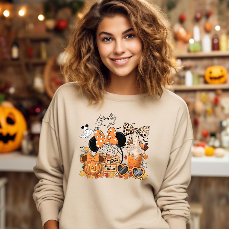 May include: Beige sweatshirt with a Halloween-themed graphic. The graphic includes a Minnie Mouse design with a pumpkin, ghost, spiderwebs, and the text "Literally just a girl". The design also features a bow, sunglasses, and fall leaves.