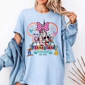 May include: Light blue t-shirt with a colorful graphic of Mickey Mouse and friends in front of the Disneyland castle. The text "Disneyland" is written in a colorful font with the tagline "The happiest place on earth -EST. 1955-."