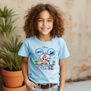 May include: A light blue t-shirt featuring a Disney Cruise design. The graphic includes Mickey Mouse, Minnie Mouse, and other characters, along with the text "Cruise" and "2025". The design is set against a backdrop of palm trees and ocean waves.