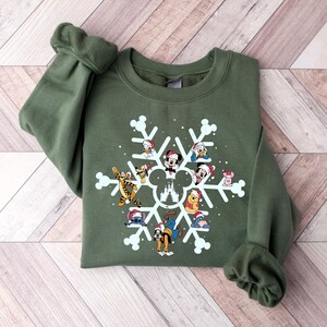 May include: A green sweatshirt with a white snowflake design featuring various Disney characters wearing Santa hats. The characters include Mickey Mouse, Minnie Mouse, Donald Duck, Goofy, Pluto, Stitch, Winnie the Pooh, and Tigger. The snowflake design also includes a silhouette of the Cinderella Castle.