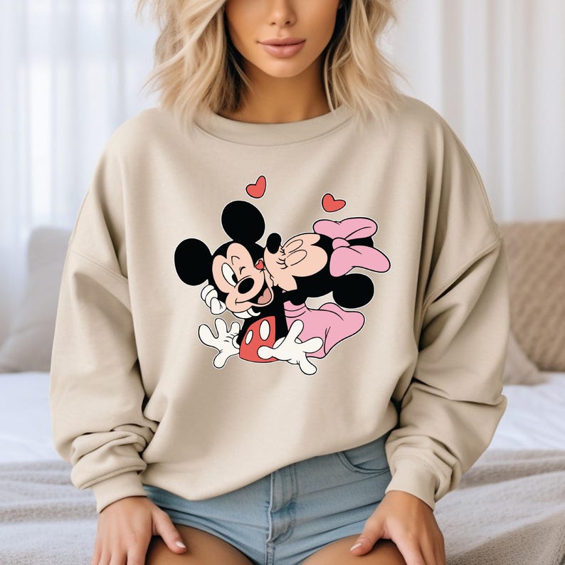May include: Beige sweatshirt featuring a graphic of Mickey Mouse kissing Minnie Mouse, with red heart accents. Mickey is wearing red shorts, and Minnie is in a pink dress and bow. The sweatshirt is a casual, comfortable style.