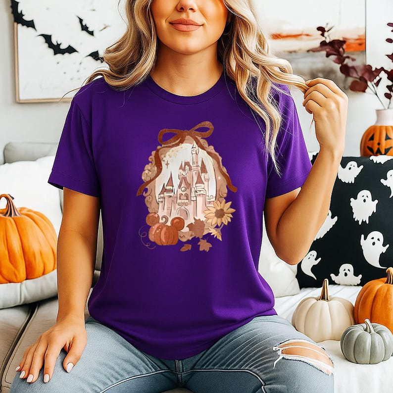 May include: A purple t-shirt with a castle design, pumpkins, sunflowers, and autumn leaves, framed by a brown ribbon. The shirt is worn by a person in blue jeans.