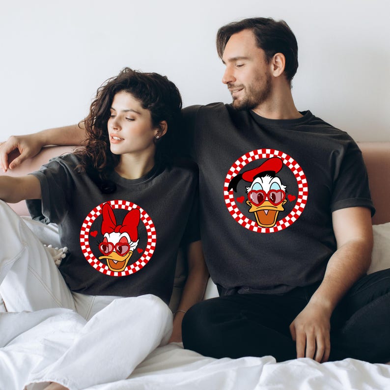 May include: A couple wearing matching black t-shirts with a red and white checkered circle design featuring a cartoon duck with red heart-shaped sunglasses. The duck is wearing a red beret.