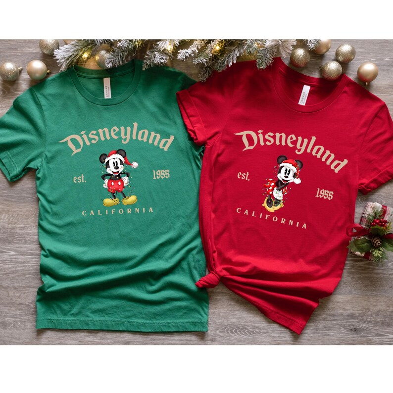 May include: Two t-shirts with a Christmas-themed Disneyland design. The green t-shirt features Mickey Mouse wearing a Santa hat and the red t-shirt features Mickey Mouse wearing a Santa hat and a red and white scarf. Both shirts have the text "Disneyland est. 1955 CALIFORNIA" printed on them.