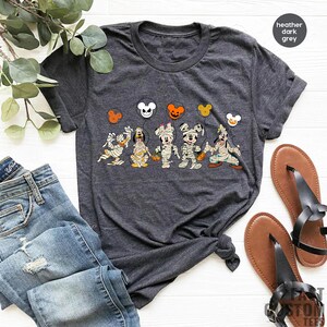 Peut inclure: Un t-shirt gris fonc&eacute; chin&eacute; avec un motif de Mickey Mouse, Minnie Mouse, Pluto et Dingo habill&eacute;s en momies tenant des ballons. Les ballons sont orange, rouge et blanc avec des oreilles de Mickey Mouse.