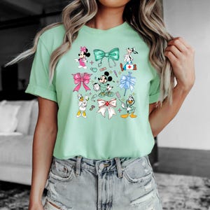 Pu&ograve; includere: T-shirt verde chiaro con personaggi dei cartoni animati e fiocchi. Il design include Minnie Mouse, Mickey Mouse, Paperino e Pippo, insieme a elementi a tema medico come uno stetoscopio e una siringa. La maglietta ha girocollo e maniche corte.