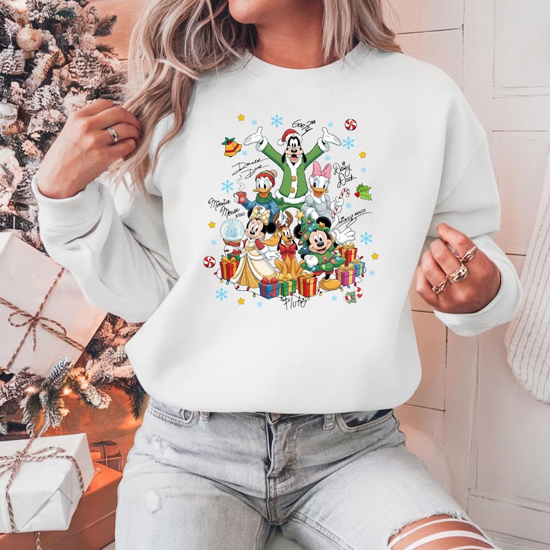 K&ouml;nnte beinhalten: Wei&szlig;es Sweatshirt mit einem festlichen Disney-Weihnachtsdesign. Die Grafik zeigt Mickey, Minnie, Donald, Daisy, Goofy und Pluto, umgeben von Schneeflocken, Geschenken und Zuckerstangen. Die Charaktere sind in Weihnachtsoutfits gekleidet.