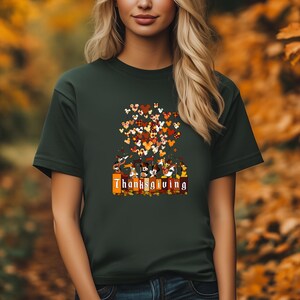 May include: Dark green t-shirt featuring a Thanksgiving design. The design includes cartoon characters, Mickey Mouse ears, and the word "Thanksgiving" in a decorative font. The design incorporates fall colors like orange, brown, and red.