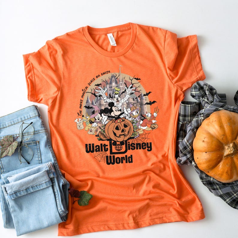Puede incluir: Camiseta naranja con un dise&ntilde;o de Halloween con personajes de Disney, una calabaza tallada y el texto "Walt Disney World". El dise&ntilde;o incluye murci&eacute;lagos, un castillo embrujado y la frase "the most magical place on earth".