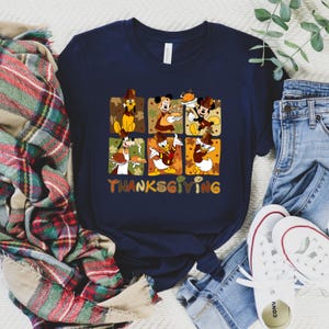 May include: Navy blue t-shirt featuring a Thanksgiving design with cartoon characters. The design includes images of Mickey Mouse, Donald Duck, and Goofy, with the word "Thanksgiving" in colorful letters. The shirt is paired with jeans and white sneakers.