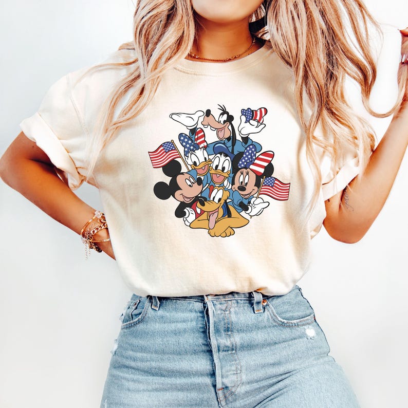 May include: Cream-colored t-shirt featuring a graphic of Mickey Mouse, Donald Duck, Goofy, and Pluto, each holding American flags. The characters are in patriotic attire, with red, white, and blue accents. The shirt is a casual style.