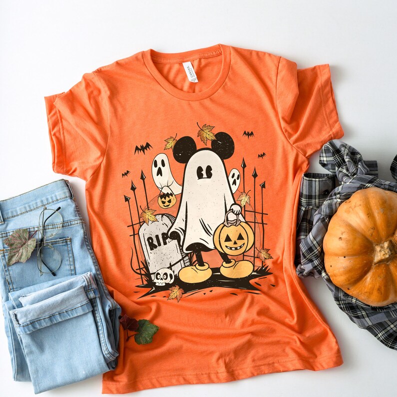 May include: Orange t-shirt with a graphic of a ghost Mickey Mouse holding a jack-o-lantern. The ghost is wearing Mickey Mouse ears and is standing in front of a graveyard with a RIP tombstone.