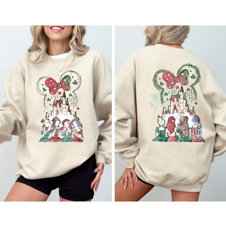 May include: Beige sweatshirt featuring a whimsical design of a castle, bow, and characters. The front and back of the sweatshirt showcase the same design. The design includes red, green, and white accents, creating a festive and charming aesthetic.