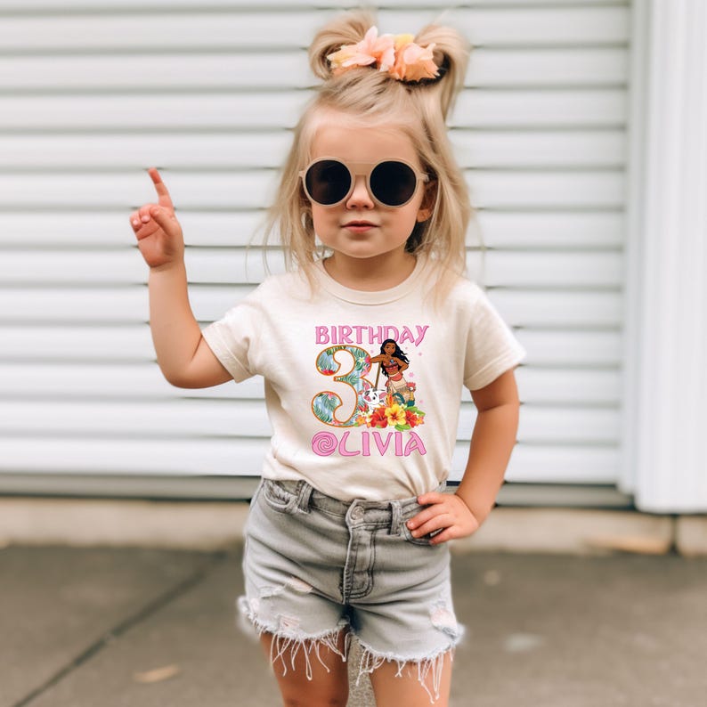 以下が含まれることがあります： 3歳の誕生日のためのカラフルなグラフィックデザインが施されたベージュのTシャツ。デザインには「BIRTHDAY」の文字、「3」の数字、「OLIVIA」の名前、花を持つ漫画のキャラクターが含まれています。子供はサングラスとグレーのショートパンツを着用しています。