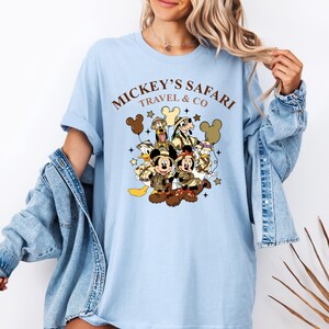 K&ouml;nnte beinhalten: Hellblaues T-Shirt mit einem Aufdruck von Mickey Mouse, Minnie Mouse, Donald Duck, Goofy und Pluto in Safari-Outfits. Der Text "Mickey's Safari Travel & Co" ist auf dem Shirt gedruckt.
