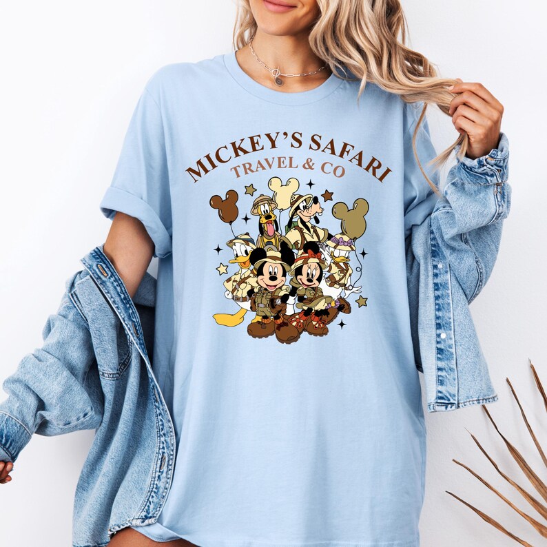 K&ouml;nnte beinhalten: Hellblaues T-Shirt mit einem Aufdruck von Mickey Mouse, Minnie Mouse, Donald Duck, Goofy und Pluto in Safari-Outfits. Der Text "Mickey's Safari Travel & Co" ist auf dem Shirt gedruckt.