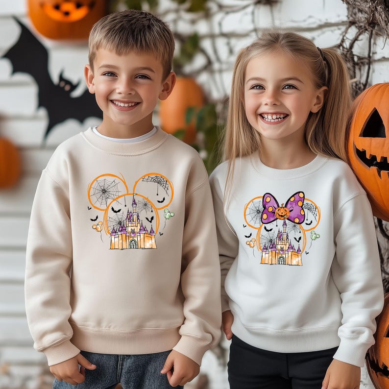 May include: Two children wearing Halloween-themed sweatshirts. One sweatshirt is beige with a castle design inside a Mickey Mouse head outline, the other is white with a bow and pumpkin design. Pumpkins and a bat silhouette are in the background.