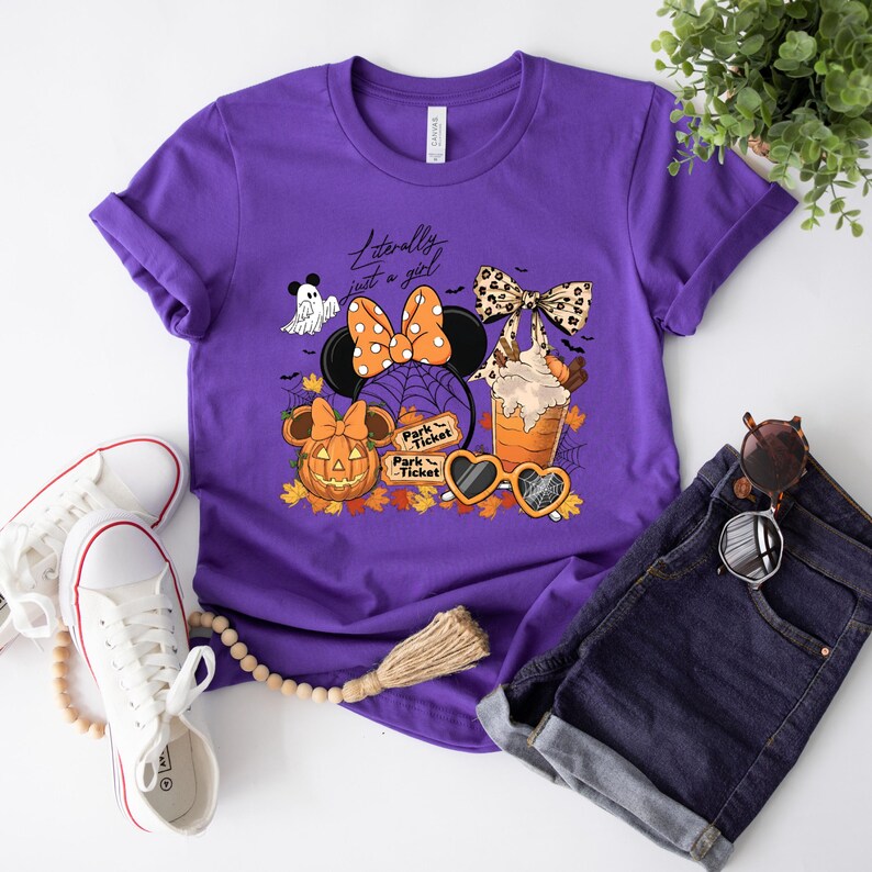 May include: Purple t-shirt with a Halloween-themed graphic featuring Minnie Mouse ears, a pumpkin, a ghost, and a sundae. The text on the shirt reads "Literally just a girl." The shirt is paired with denim shorts and white sneakers.