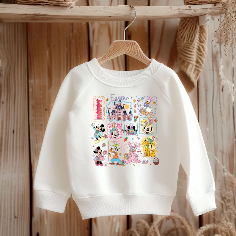 May include: White sweatshirt featuring a colorful Easter-themed graphic with Disney characters. The design includes a castle, bunnies, and the words "Happy" and "Easter". The sweatshirt is displayed on a wooden hanger.
