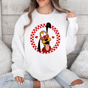 May include: White sweatshirt with a red and white checkered circle design featuring a cartoon dog wearing heart-shaped sunglasses. The dog is sticking its tongue out.