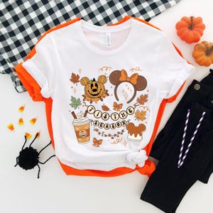 May include: White t-shirt with an orange trim featuring a fall-themed graphic. The design includes a pumpkin, Mickey Mouse ears, fall leaves, and the text "Tis the Season". The shirt is paired with black pants and decorative pumpkins.