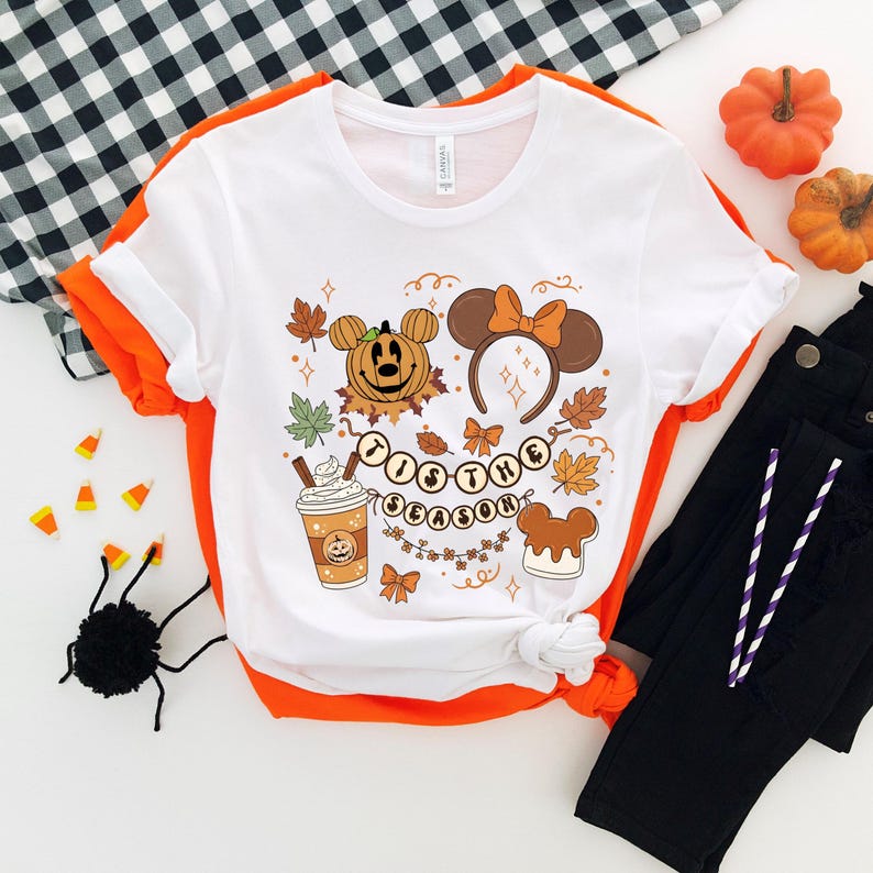 May include: White t-shirt with an orange trim featuring a fall-themed graphic. The design includes a pumpkin, Mickey Mouse ears, fall leaves, and the text "Tis the Season". The shirt is paired with black pants and decorative pumpkins.