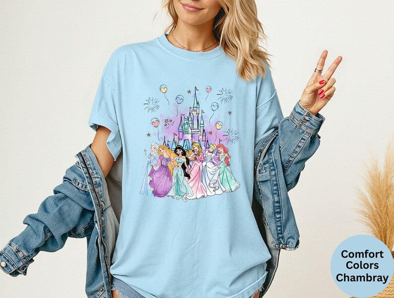 K&ouml;nnte beinhalten: Hellblaues T-Shirt mit einer Aquarellillustration eines Schlosses und Prinzessinnen. Das Design umfasst Feuerwerk und Ballons. Das Shirt ist mit "Comfort Colors Chambray" gekennzeichnet.