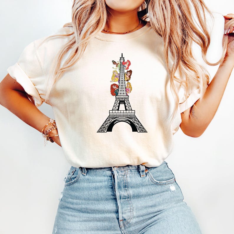 May include: Cream-colored t-shirt featuring a black Eiffel Tower design with colorful cartoon princess figures. The graphic is centered on the front of the shirt. The shirt has short sleeves and a crew neck.