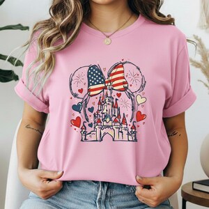 May include: Pink t-shirt featuring a graphic of a castle with an American flag bow and mouse ears. The design includes red, white, and blue hearts and fireworks. The shirt is made of a soft material.