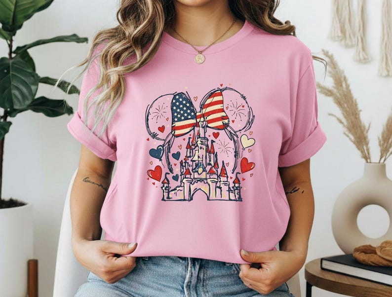 May include: Pink t-shirt featuring a graphic of a castle with an American flag bow and mouse ears. The design includes red, white, and blue hearts and fireworks. The shirt is made of a soft material.
