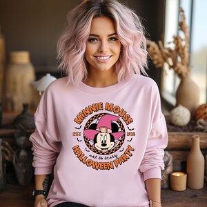 May include: Pink sweatshirt featuring a graphic of Minnie Mouse dressed as a witch. The graphic includes the text "Minnie Mouse Halloween Party Est. 1928 Not so scary".