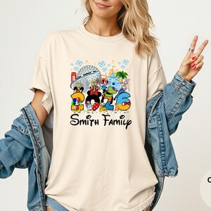 May include: Ivory-colored t-shirt featuring a colorful graphic with Disney characters and the numbers "2026". The design includes iconic park landmarks and the text "Smith Family" in a script font. The shirt is a comfort colors style.