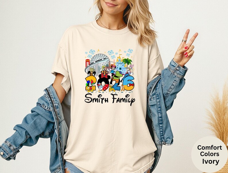 May include: Ivory-colored t-shirt featuring a colorful graphic with Disney characters and the numbers "2026". The design includes iconic park landmarks and the text "Smith Family" in a script font. The shirt is a comfort colors style.