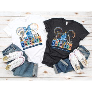 May include: Two t-shirts with a graduation theme featuring Mickey Mouse and friends at Disney World. The white t-shirt has the year 2025 printed on it. Both shirts have the text "Graduate from the tassel to the castle".